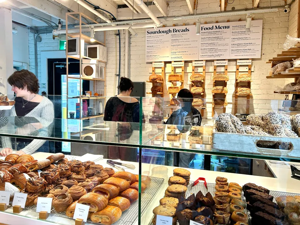A bakery counter filled with fresh pastries, croissants, cookies, and breads, with staff working behind glass partitions and a menu board on the wall.