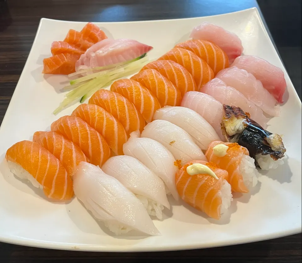 A large white platter filled with an assortment of sushi nigiri, including salmon, white tuna, and unagi, alongside fresh salmon and white fish sashimi with cucumber garnish.
