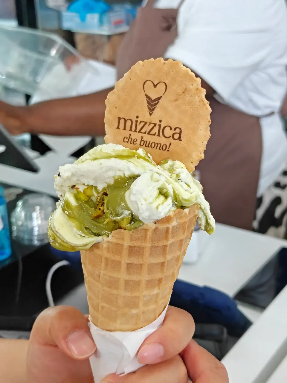 A hand holding a waffle cone filled with pistachio and cream gelato, topped with a round wafer cookie featuring the Mizzica logo.