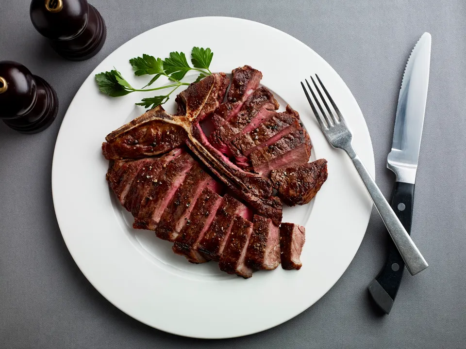 A sliced T-bone steak served on a white plate with a sprig of parsley, accompanied by a fork, knife, and pepper grinders on a gray surface.