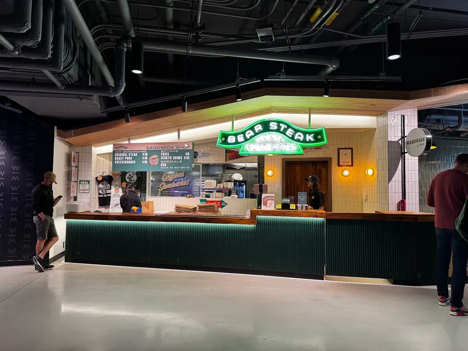 A sandwich shop counter with a bright green neon sign that reads 'BEAR STEAK', a menu board, and staff serving customers. Exposed industrial pipes are visible on the ceiling.