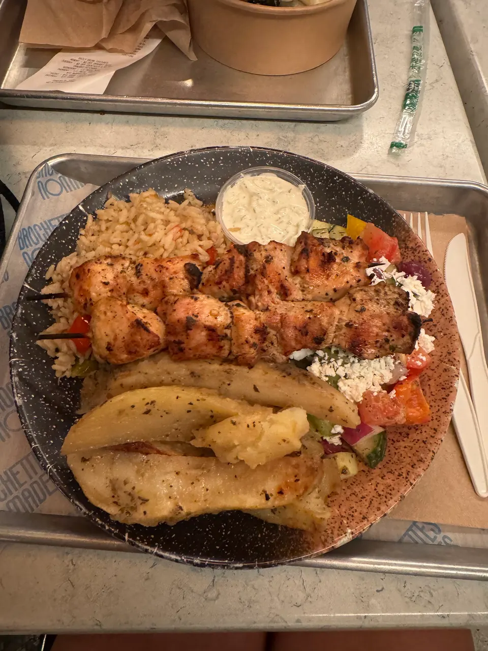 A Greek platter on a speckled plate featuring two grilled chicken skewers, seasoned rice, roasted potato wedges, a side salad with feta cheese, and a small cup of tzatziki sauce.