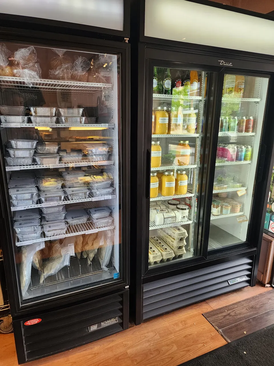 Two commercial glass-door refrigerators at Dough Bakeshop, stocked with packaged prepared meals, baked goods, bottled juices, and other refrigerated items.