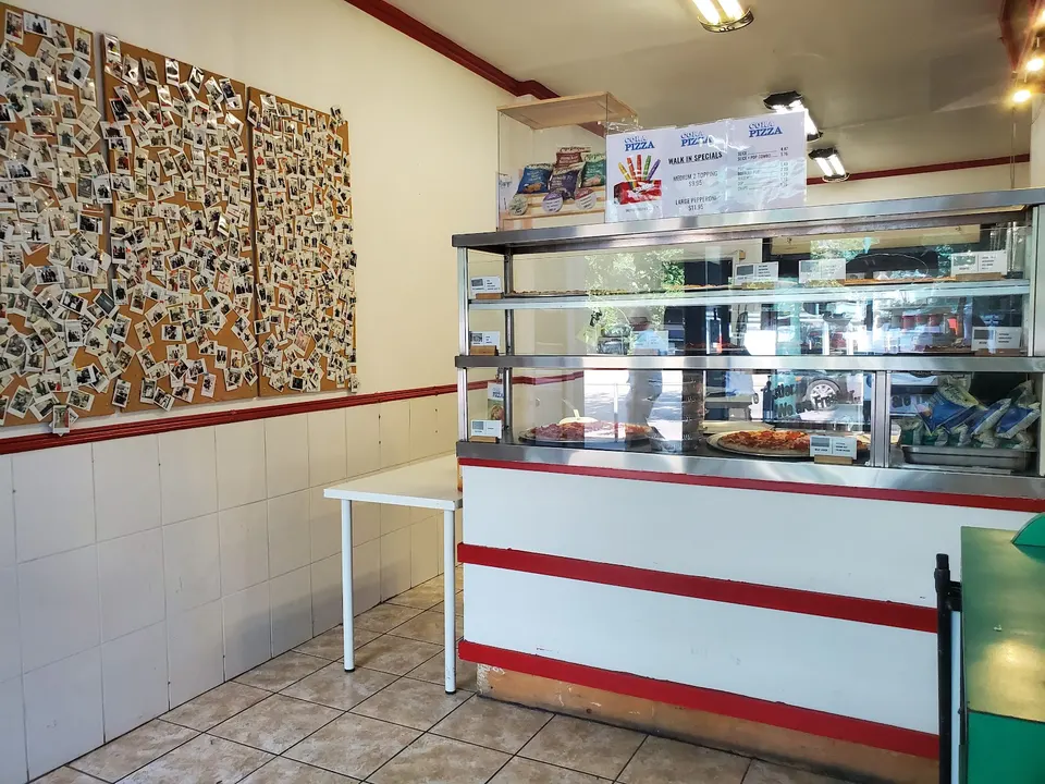 Interior of Cora Pizza showing a glass display counter with pizzas, a white table, and a large corkboard covered in polaroid photos on a white tiled wall.