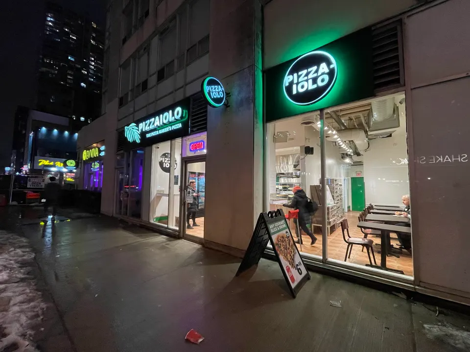 Exterior view of Pizzaiolo at night, showing the bright green neon signs, a large window revealing the indoor dining area with tables and chairs, and a sidewalk A-frame sign outside.