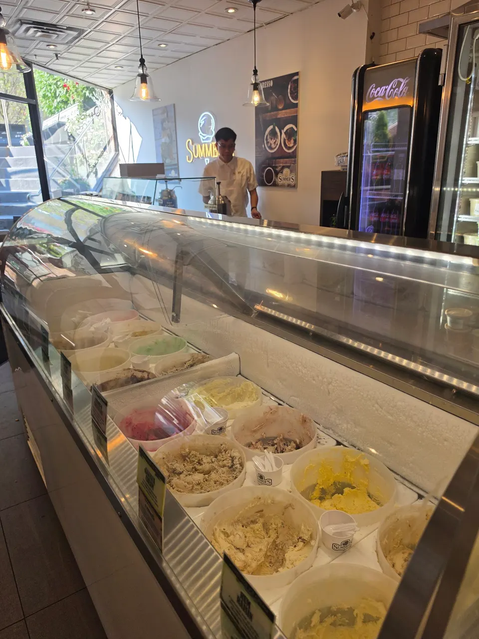 A glass display case filled with various tubs of ice cream flavors at Summer's Ice Cream, with a staff member standing behind the counter.