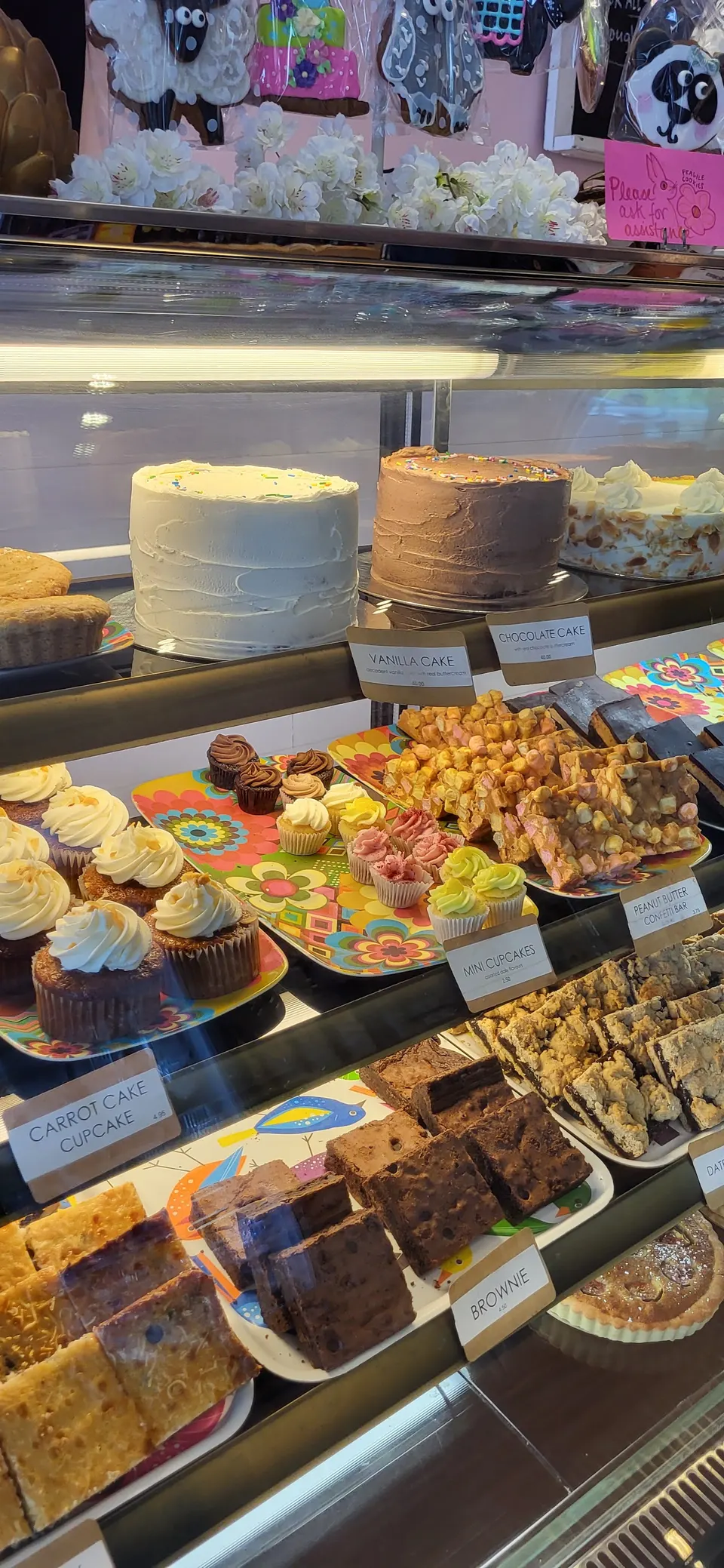 A bakery display case filled with various desserts including whole cakes, cupcakes, brownies, and bar treats with handwritten labels.