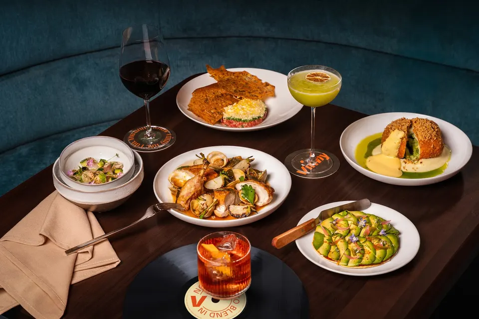 A dark wooden table at Vinny Toronto featuring a spread of Italian-inspired dishes, including sliced avocado, roasted chicken, and a breaded main, alongside cocktails and a glass of red wine.