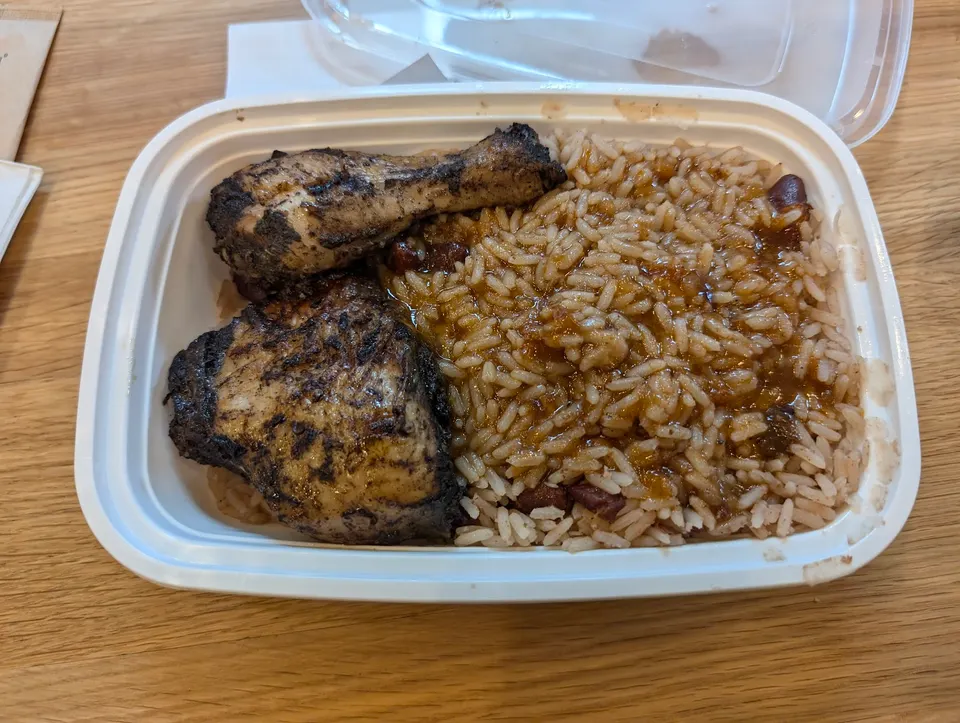 A white rectangular takeout container filled with two pieces of grilled jerk chicken and a generous portion of rice and peas. The chicken pieces are dark brown with visible grill marks, and the rice is light brown with red kidney beans mixed in, coated in a savory sauce. The container sits on a ligh