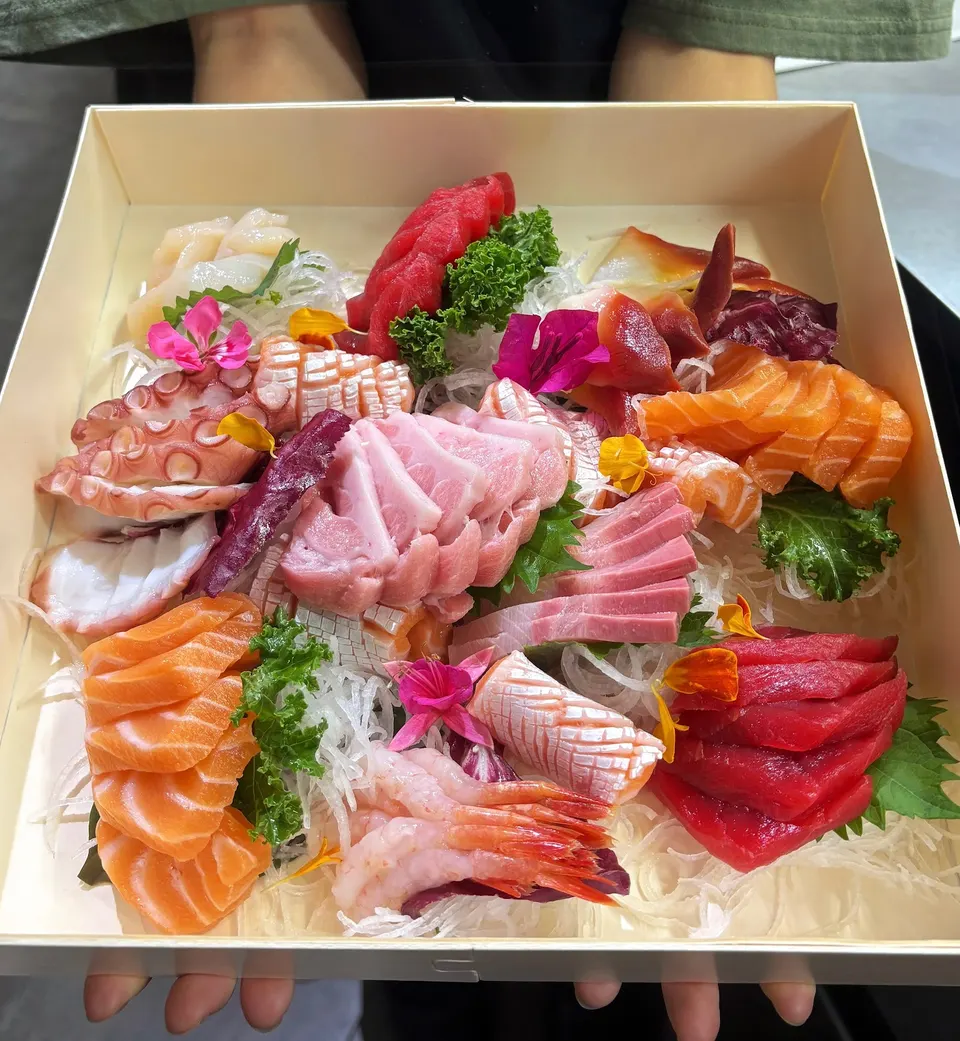 A wooden box filled with an assortment of fresh sashimi, including salmon, tuna, octopus, and sweet shrimp, beautifully arranged with edible flowers and shredded daikon radish.