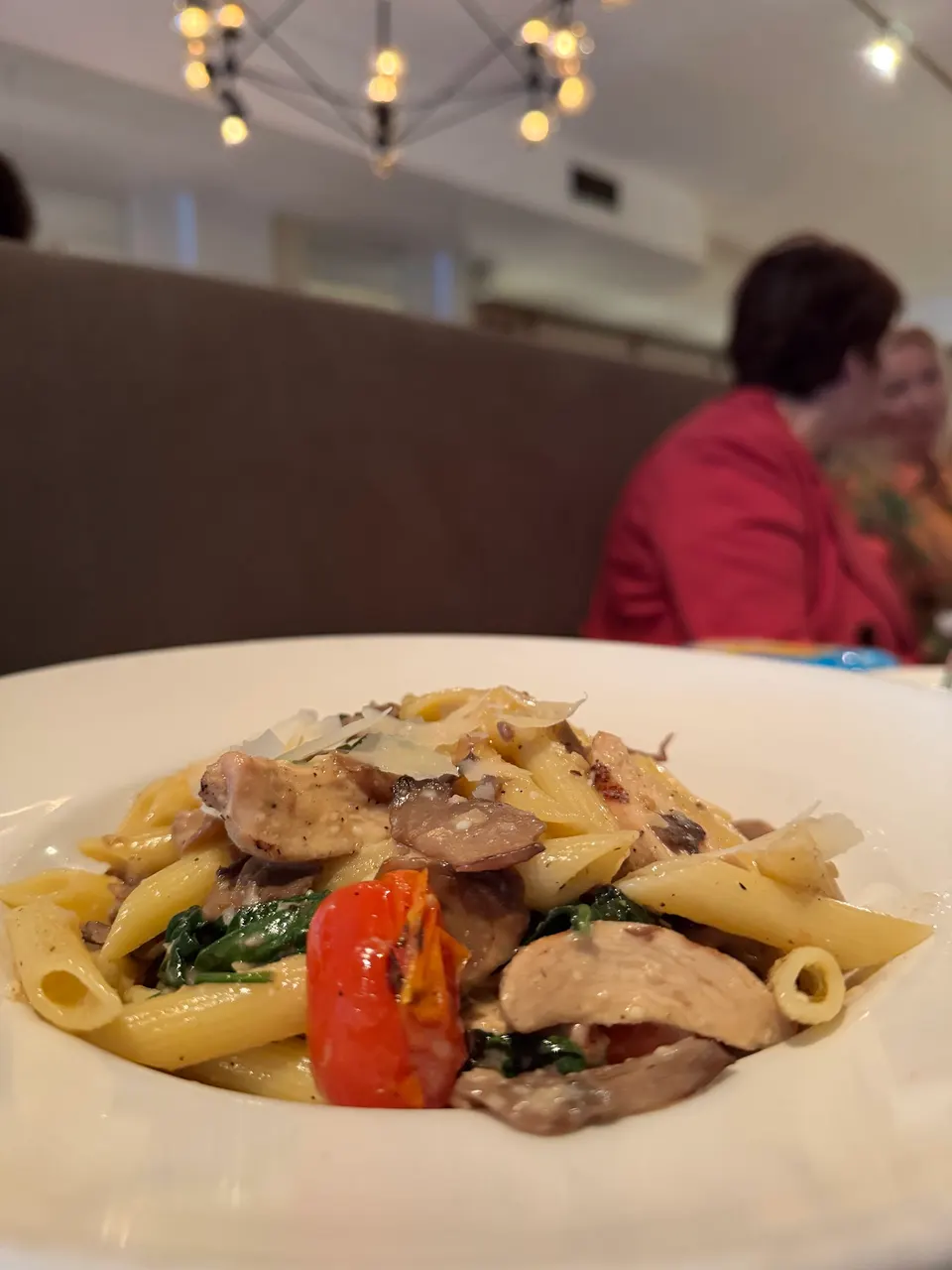 A close-up shot of a penne pasta dish served in a white bowl, featuring mushrooms, spinach, cherry tomatoes, chicken, and shaved parmesan cheese, with a blurred restaurant background.