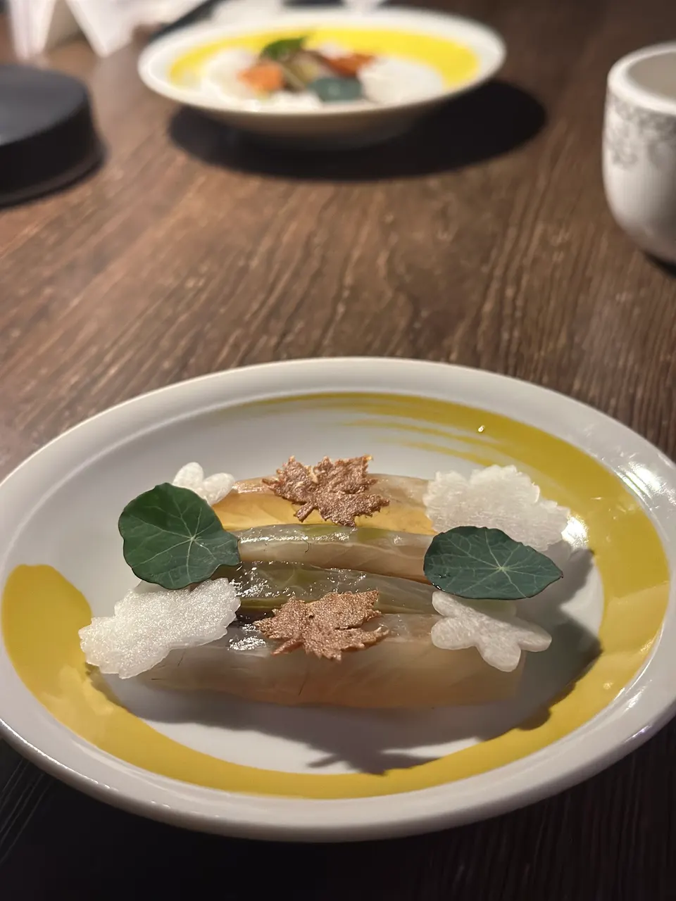 A beautifully plated dish at Yan Dining Room featuring delicate slices of fish garnished with leaf-shaped crisps, white radish cutouts, and green nasturtium leaves on a yellow-rimmed plate.