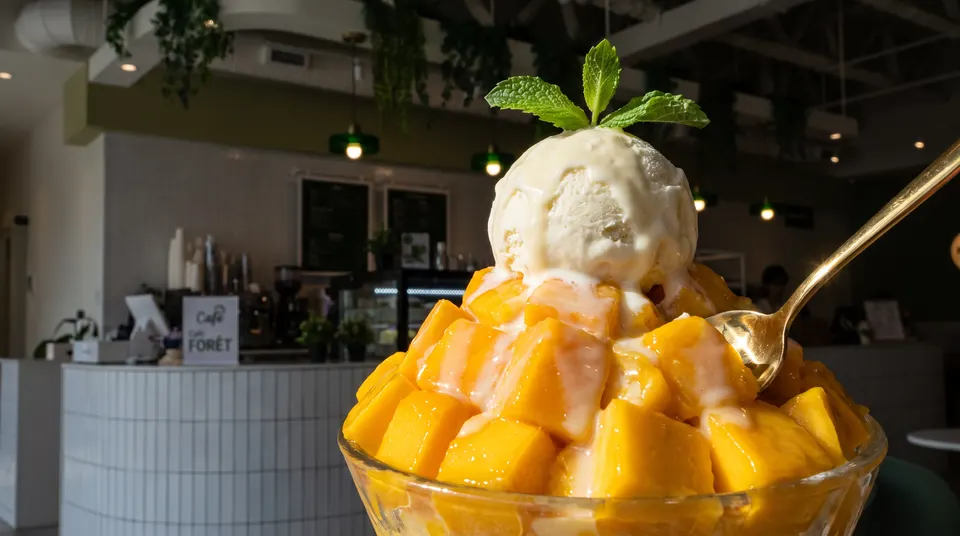 A large bowl of mango bingsu topped with a scoop of vanilla ice cream, condensed milk, and a mint sprig, served with a gold spoon at Cafe Forêt.