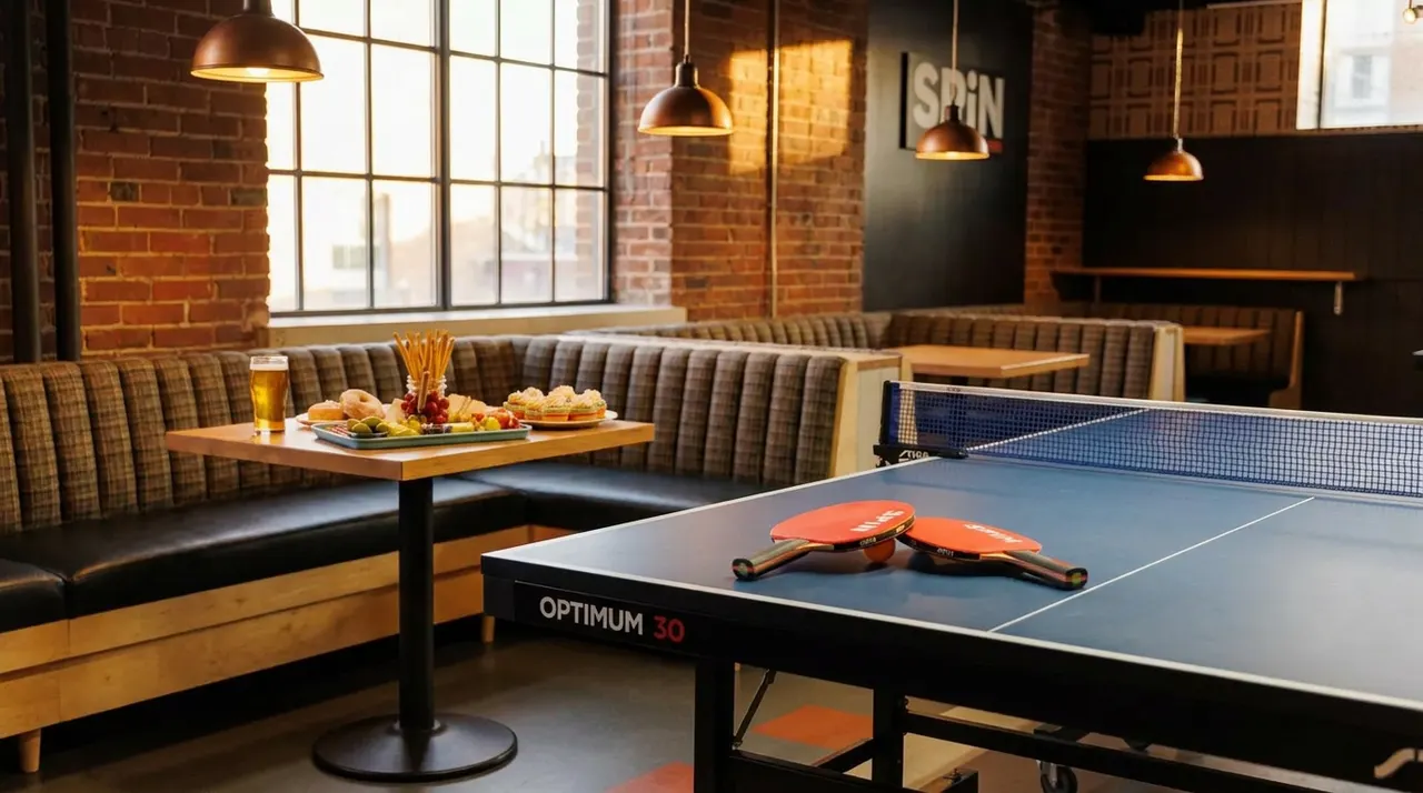 A blue ping pong table with two paddles sits in the foreground of a cozy bar lounge featuring brick walls, leather booth seating, and a wooden table set with snacks and a beer.