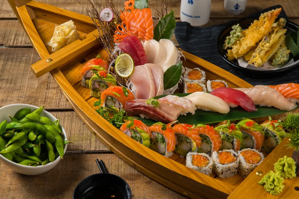 A wooden sushi boat filled with an assortment of sushi rolls, nigiri, and fresh sashimi, served alongside edamame, tempura, and wasabi on a rustic wooden table.
