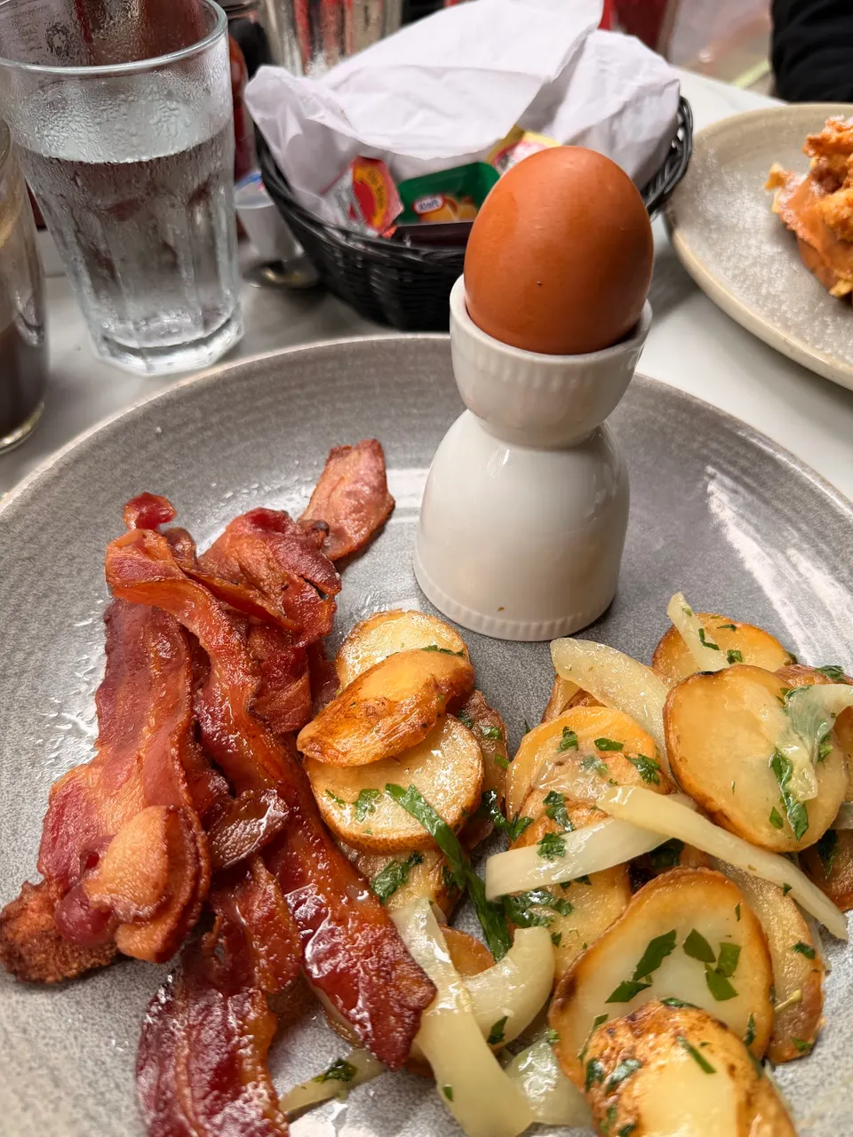 A breakfast plate featuring crispy bacon strips, roasted sliced potatoes with herbs and onions, and a soft-boiled brown egg served in a white ceramic egg cup.