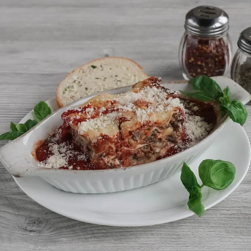 A slice of layered lasagna topped with grated parmesan cheese and marinara sauce, served in an oval white baking dish with a side of garlic bread and fresh basil leaves.