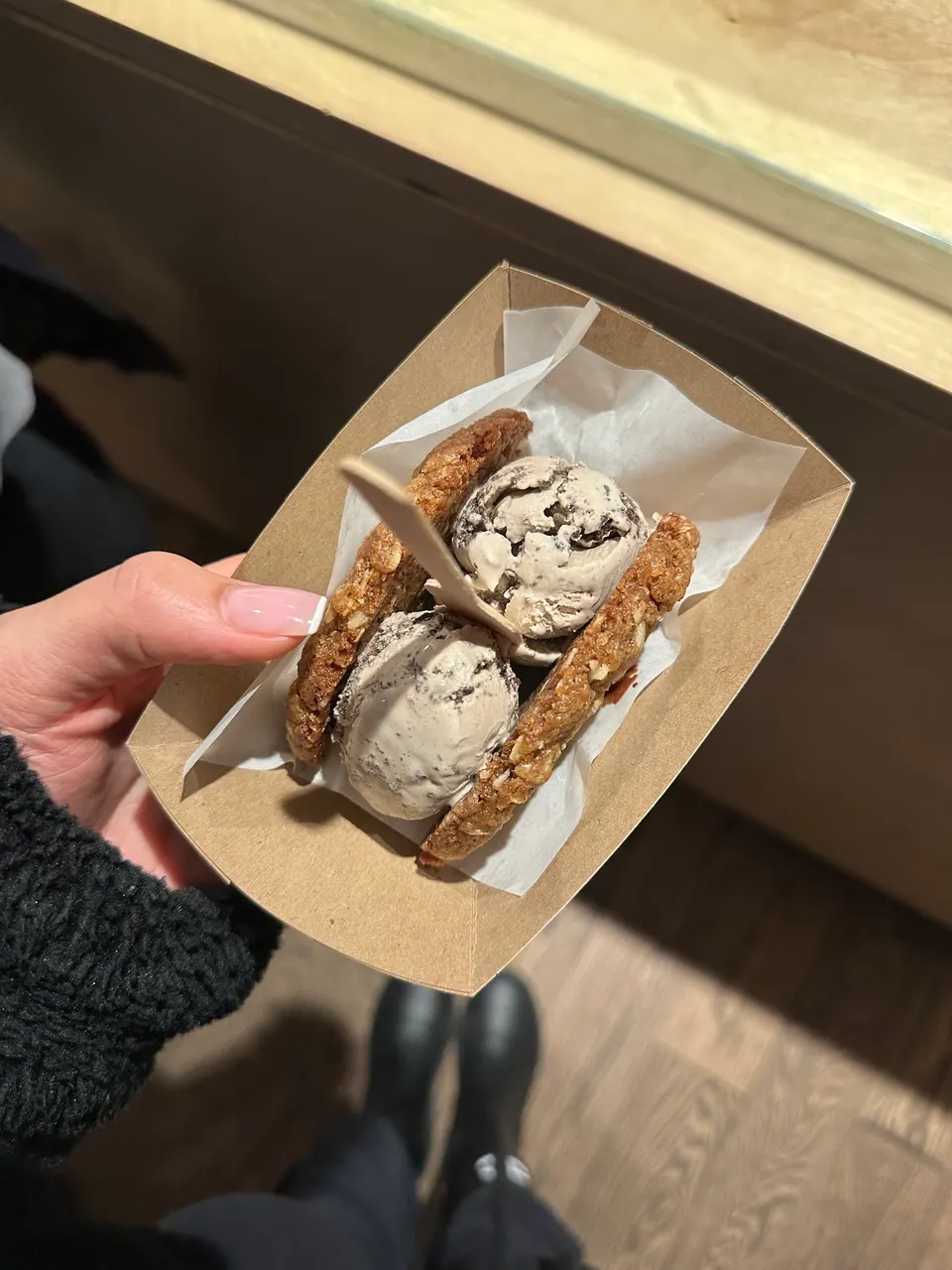 A hand holding a cardboard tray with an ice cream sandwich made of two oatmeal cookies and two scoops of cookies and cream ice cream, with a small wooden spoon inserted.