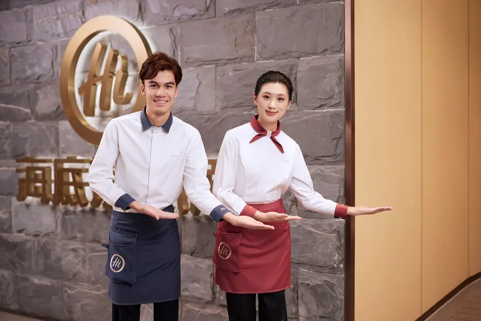 A male and female server in clean white uniforms with aprons smile and gesture welcomingly in front of a stone wall featuring the golden Haidilao logo.