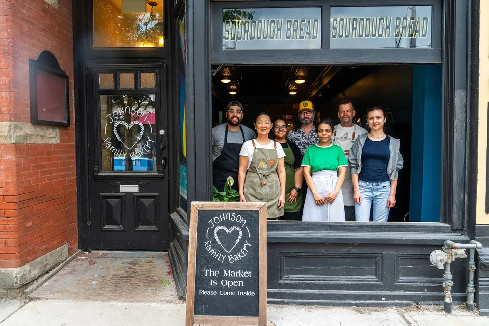 The staff of Johnson Family Bakery standing together inside the front window of their storefront, with a chalkboard sign on the sidewalk outside.