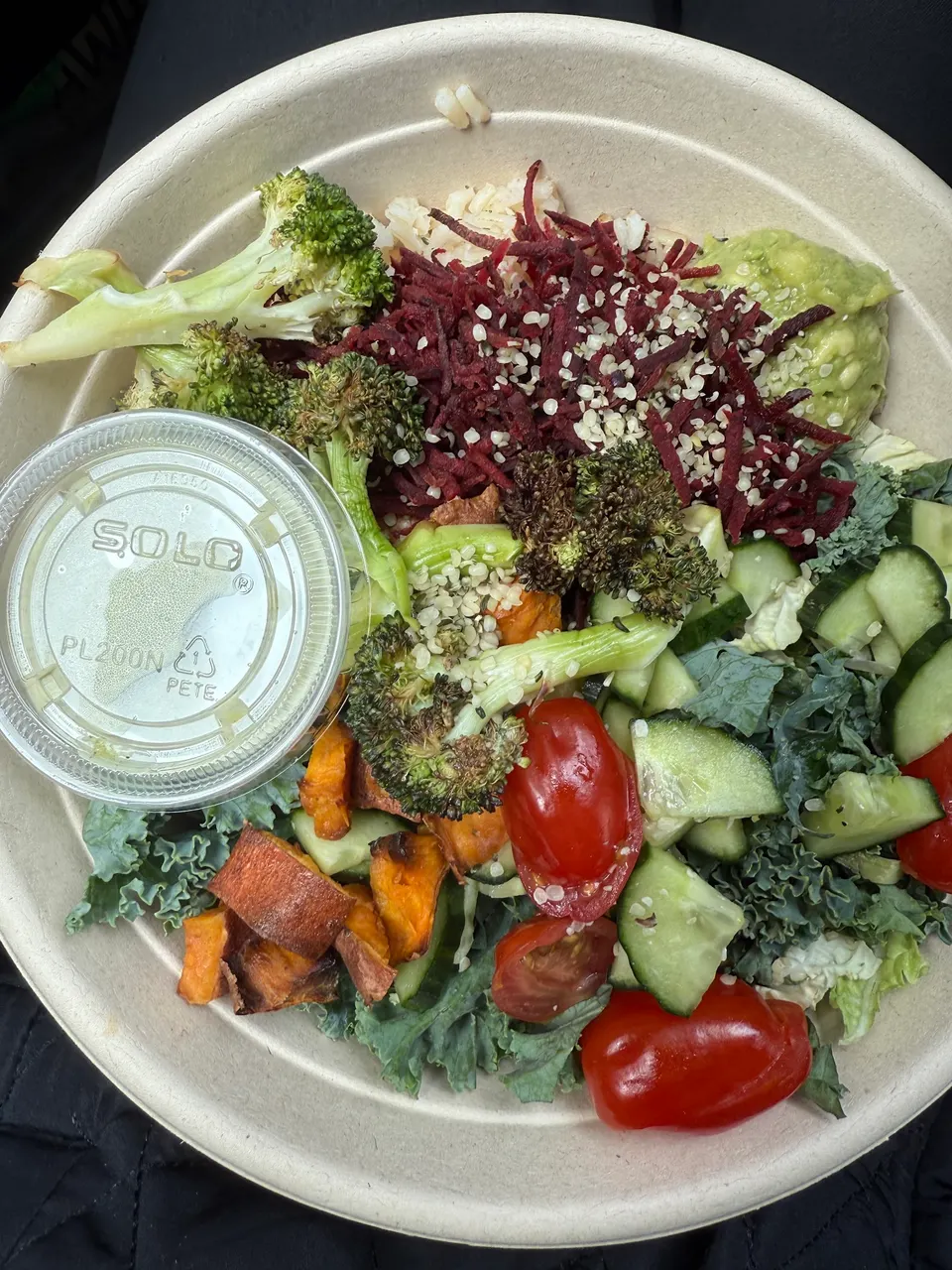 A healthy salad bowl from Salus Fresh Foods containing kale, cucumbers, cherry tomatoes, roasted sweet potatoes, broccoli, shredded beets, hemp seeds, guacamole, and a side of dressing in a plastic cup.
