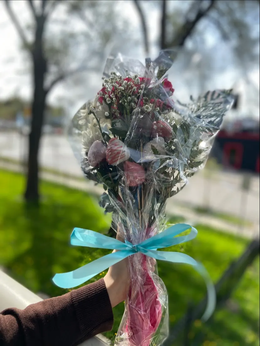 A hand holding a bouquet of fresh flowers mixed with chocolate-covered strawberries, wrapped in clear cellophane and tied with a bright blue ribbon, set against a blurry outdoor background.