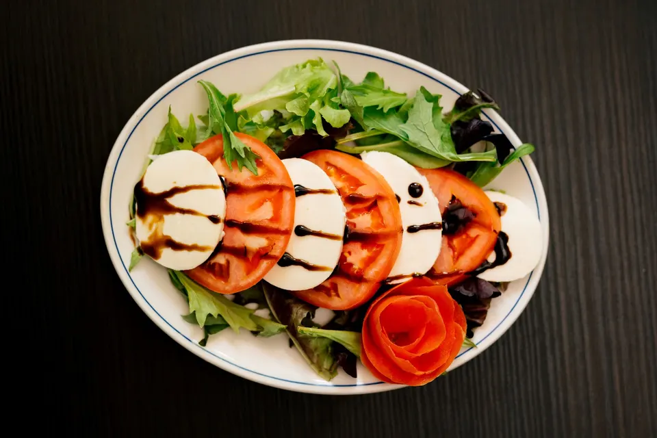 A fresh Caprese salad arranged on a bed of mixed greens in an oval white plate, featuring alternating slices of ripe red tomatoes and fresh mozzarella cheese drizzled with balsamic glaze, garnished with a tomato skin rose.