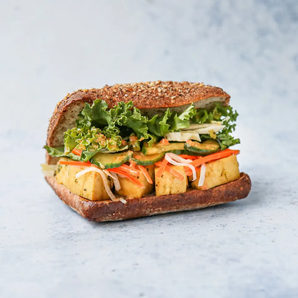 A gourmet tofu sandwich on multi-grain bread, layered with marinated tofu cubes, pickled carrots, cucumber slices, and fresh green lettuce, set against a light blue textured background.
