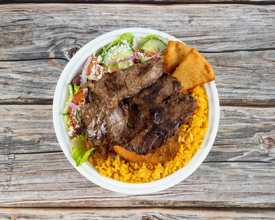 A Greek street food bowl filled with yellow rice, grilled steak slices, a side salad with feta cheese, and pita bread triangles, served in a white bowl on a rustic wooden table.