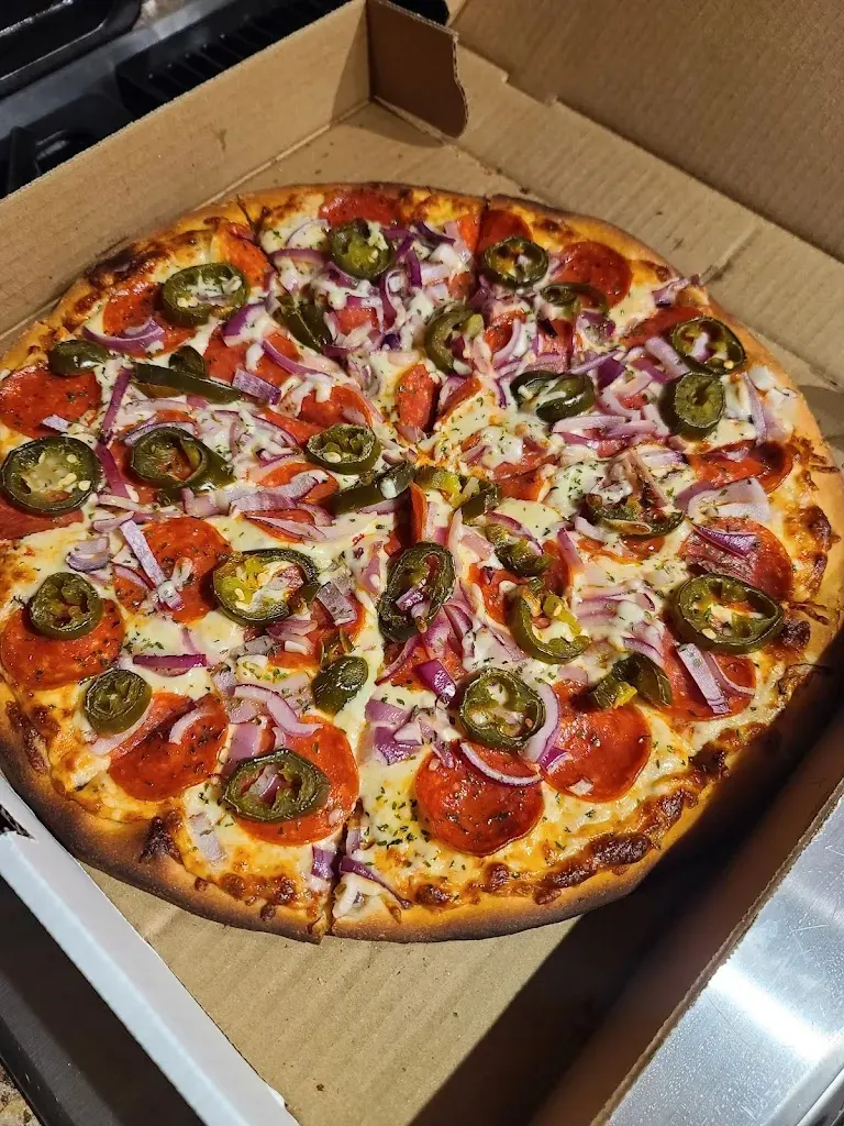 A freshly baked pizza in a cardboard delivery box, topped with melted cheese, pepperoni slices, red onions, and sliced green jalapeños.