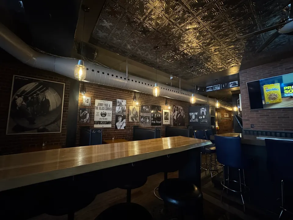 Interior of Factory Girl bar featuring a long wooden counter with bar stools, brick walls adorned with music posters, hanging pendant lights, and a textured metallic ceiling.