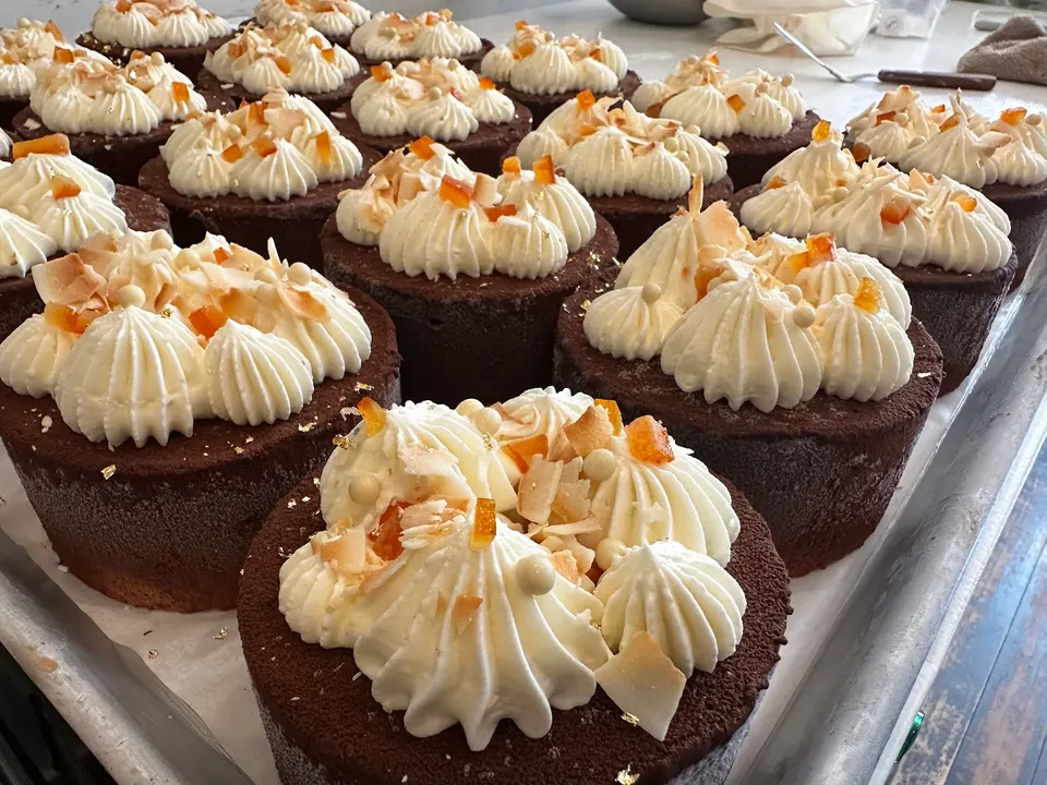 A metal baking tray filled with rows of individual round chocolate cakes, each topped with piped white frosting, toasted coconut flakes, and candied orange peel.