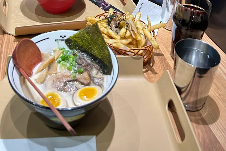 A bowl of ramen with soft-boiled egg, pork belly, green onions, and nori seaweed, served on a tray next to french fries and a metal cup of water.