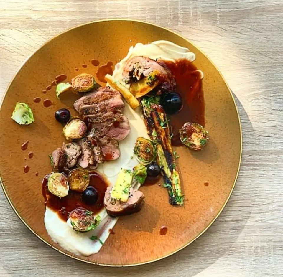 A beautifully plated gourmet dish featuring sliced roasted meat, charred vegetables, brussels sprouts, and a rich dark sauce over a creamy puree, served on a round golden plate.