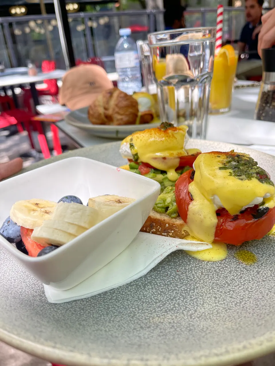 A plate of eggs benedict topped with hollandaise sauce and herbs, served alongside a small white bowl of sliced bananas and blueberries on a grey plate.