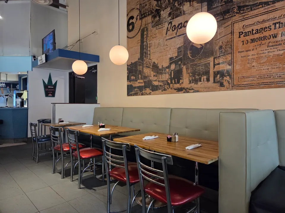 A row of wooden dining tables paired with red-cushioned metal chairs and light green booth seating, set against a wall featuring a large vintage-style mural under hanging globe lights.