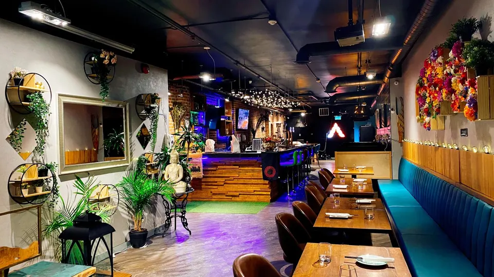 Interior of Crystal Thani Kitchen & Bar showing a long dining area with a blue banquette, wooden tables, and dark chairs, leading towards a bar counter with colorful lighting.