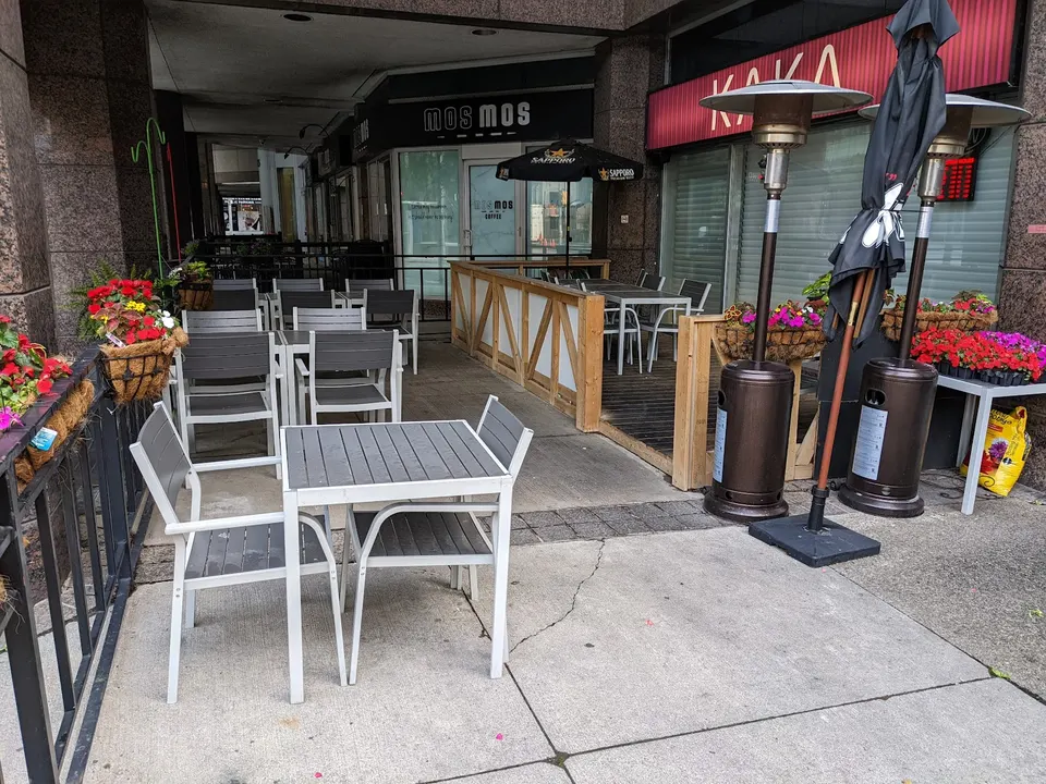 An outdoor patio dining area with grey metal tables and chairs, patio heaters, and flower boxes, situated in front of the KAKA and Mos Mos storefronts.
