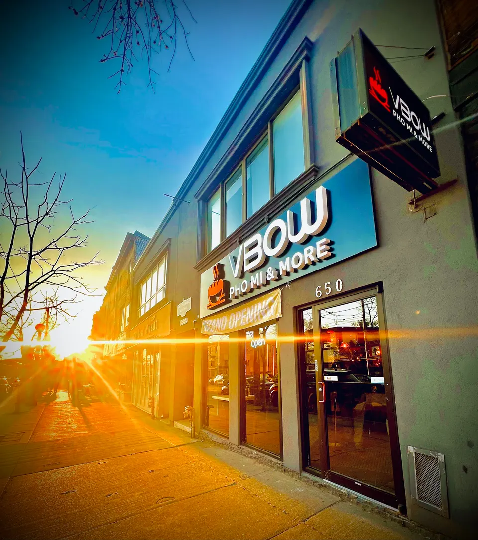 Exterior storefront of VBOW Pho Mi & More at 650, featuring a large illuminated sign and a smaller hanging sign against a sunset sky.