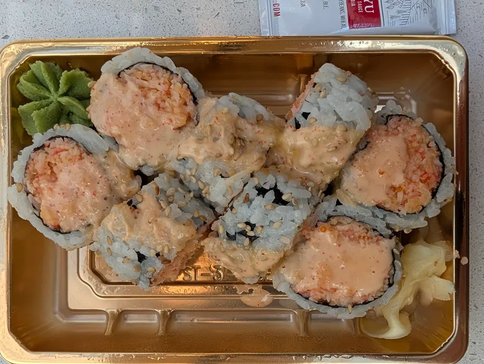 A golden takeout container filled with eight pieces of spicy crab sushi rolls topped with creamy sauce and sesame seeds, served with wasabi and pickled ginger.
