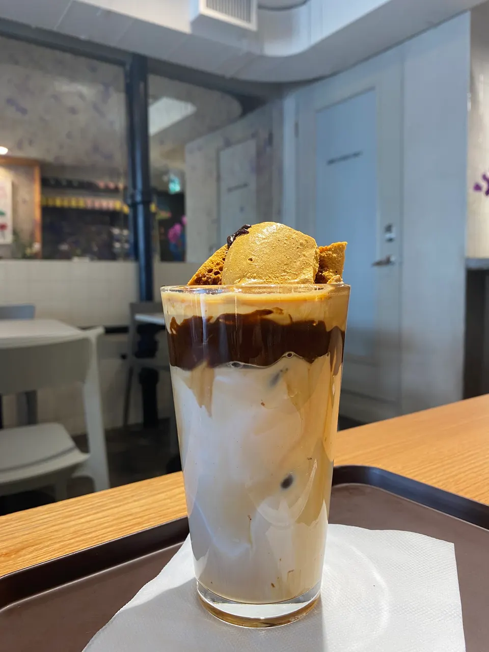 An iced coffee drink in a tall glass, layered with milk, espresso, chocolate sauce, and topped with honeycomb toffee pieces, served on a tray at Cafe Kray.
