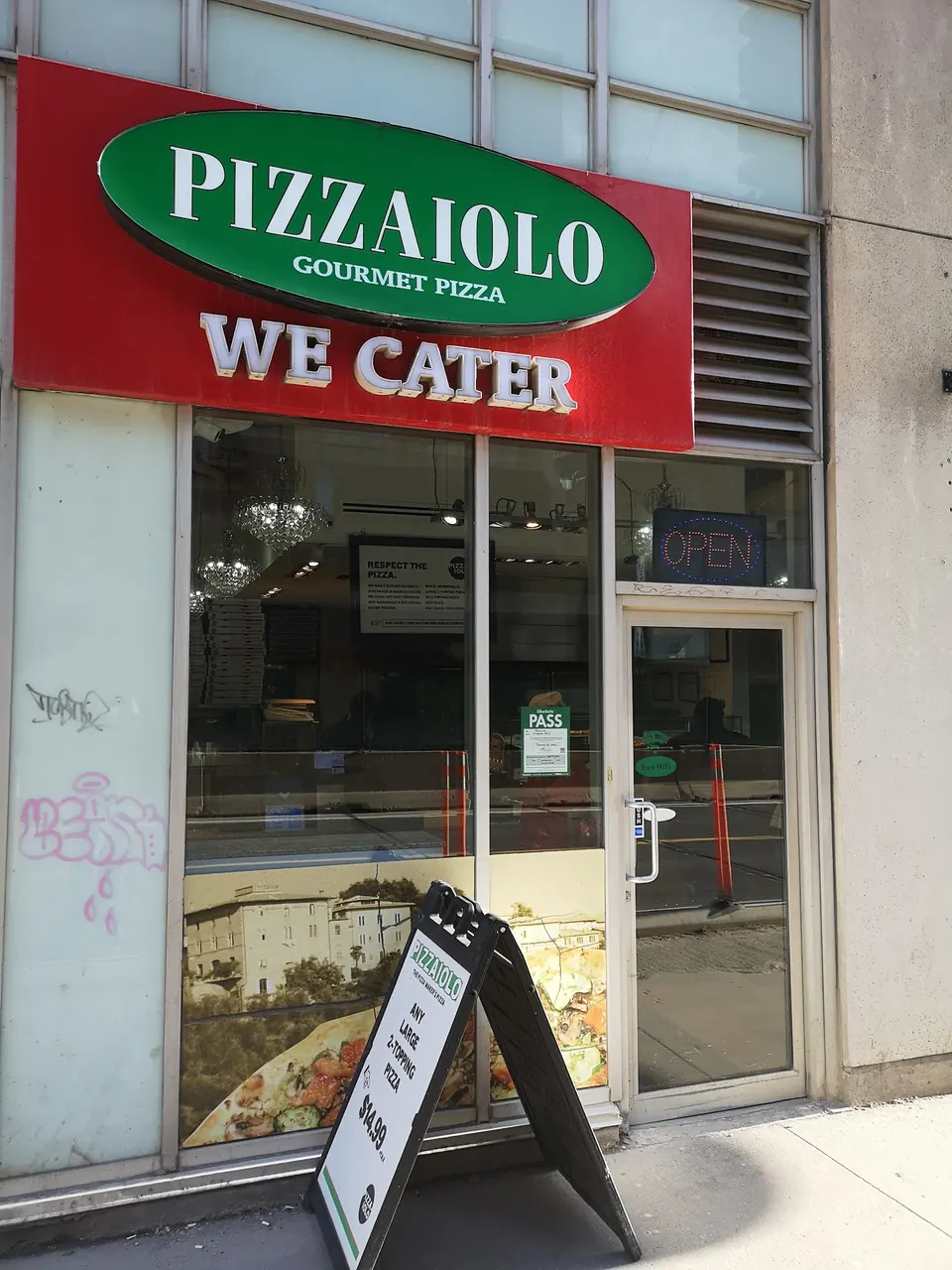 Street-level storefront of Pizzaiolo Gourmet Pizza, featuring a red and green sign, glass windows, a door, and an A-frame sidewalk sign advertising a pizza special.