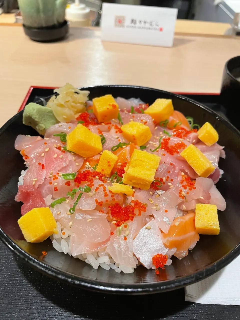 A colorful chirashi don bowl filled with assorted sashimi, cubed tamago, and red fish roe over sushi rice, served with wasabi and pickled ginger.