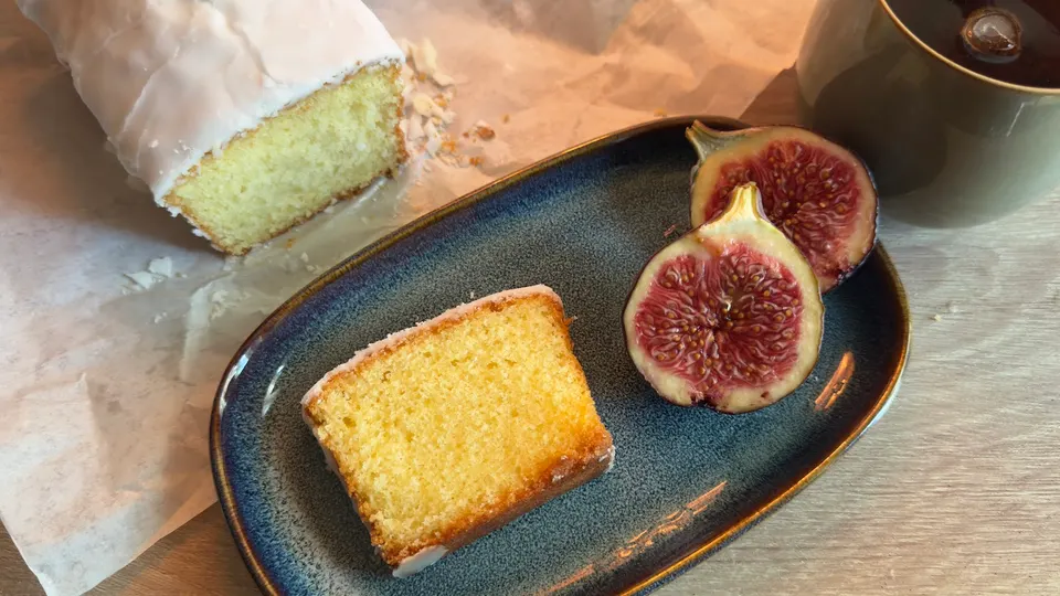 A slice of iced lemon loaf cake and two halved fresh figs served on a blue ceramic plate, with the remaining loaf on parchment paper and a mug nearby.