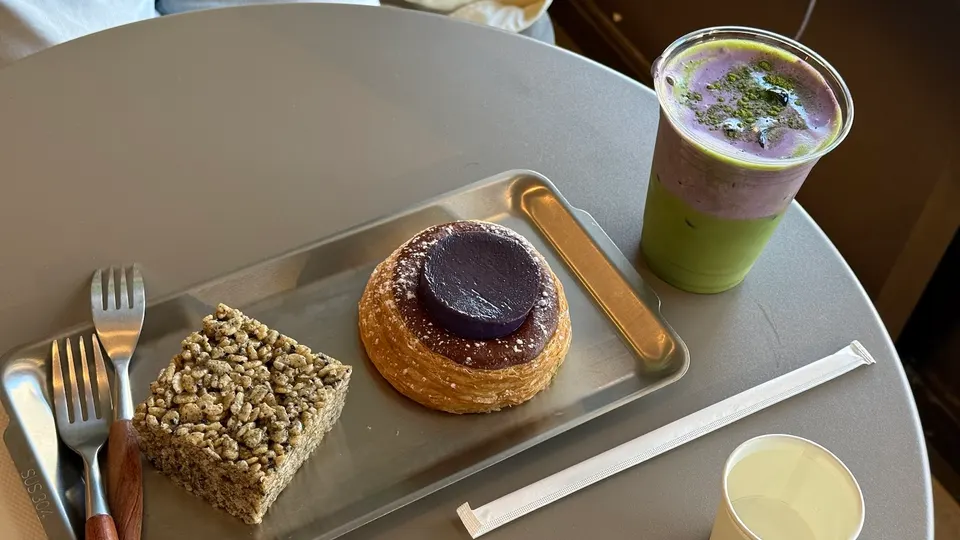 A metal tray on a round table holds a square puffed rice treat and a round pastry topped with a purple ube disc. A layered green and purple matcha drink sits nearby.