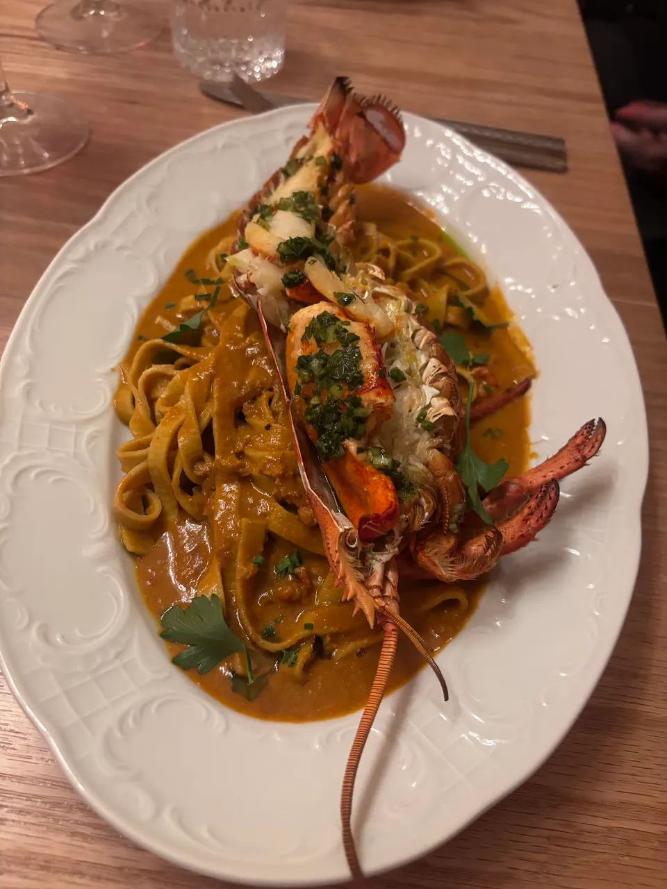 A plate of fresh tagliatelle pasta tossed in a rich orange-brown sauce, topped with a split grilled lobster tail garnished with chopped green herbs, served on an ornate white oval platter.