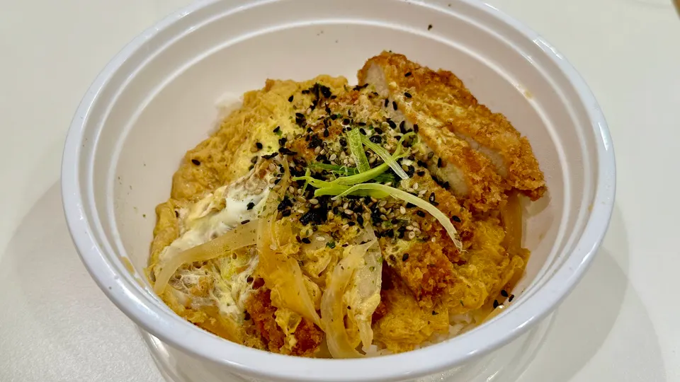 A katsu donburi bowl with breaded pork cutlet, simmered egg, onions, and green onions over rice in a white takeout bowl.