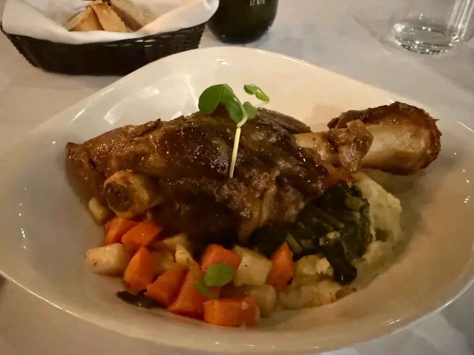 A slow-cooked lamb shank served on a bed of mashed potatoes with roasted carrots, parsnips, and greens in a white bowl.