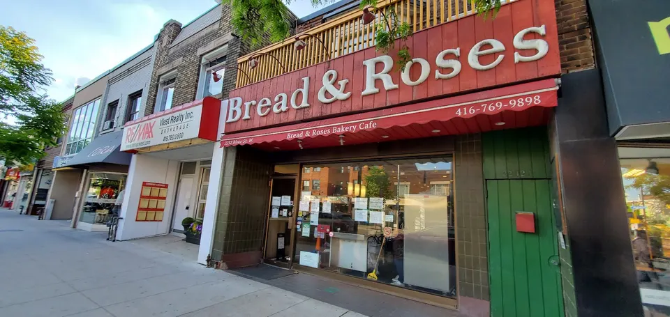 Street-level exterior view of Bread & Roses Bakery Cafe, featuring a large red wooden sign with white lettering, a red awning, and a glass storefront next to a green door.