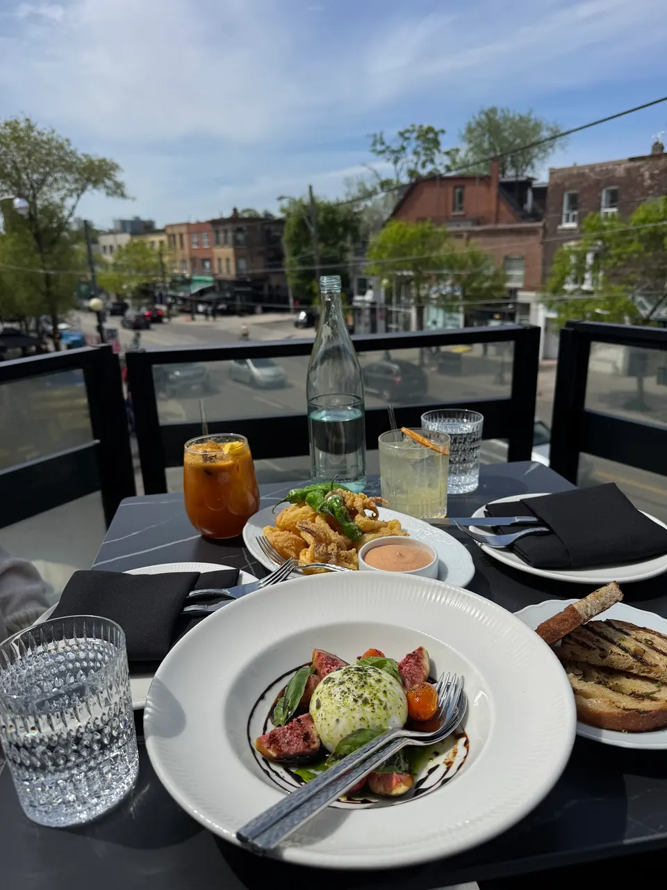 An outdoor restaurant table set with a burrata salad, fried calamari, grilled bread, cocktails, and a bottle of water, overlooking a city street from a rooftop patio.